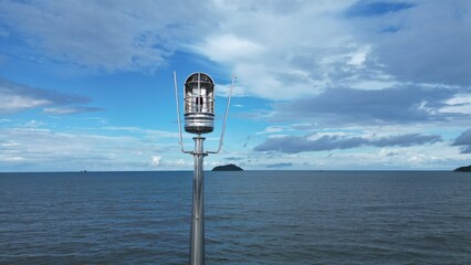 Close-up of a metallic navigation light on a pier with a vast blue sea and cloudy sky in the background.