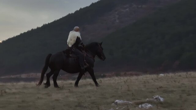 Cinematic aerial drone shot of a Viking warrior on a large black horse surveys a dramatic landscape during sunset, wearing a fur cloak concept of Ancient exploration, adventure, and the spirit of the