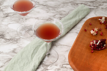 Two pomegranate cocktails in sugar rimmed martini glasses on white and gray marbled surface.