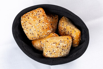 Four fresh whole-grain bread rolls covered with sesame and flax seeds, served in a black bread basket on a white tablecloth.