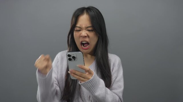 Chinese woman frustrated with phone isolated against grey background expressing anger and disappointment.