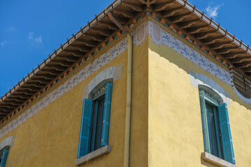 Sunny view of a yellow stucco building with teal shutters, decorative floral border patterns, and a wooden roof overhang against a clear blue sky.