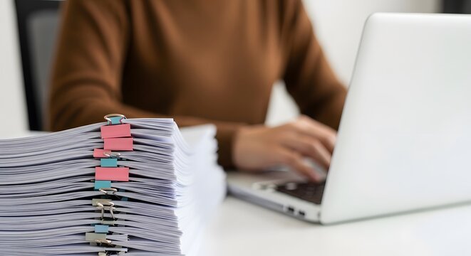 Busy employee at a desk handling a heavy workload of documents and typing on a laptop computer