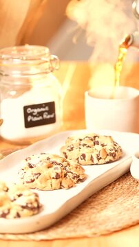 Three chocolate chip cookies on a white plate with a jar of sugar and a cup of tea. The cookies are arranged in a row and the tea is poured into the cup