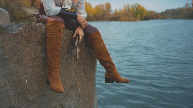 unrecognizable silhouette fantasy woman pirate armed with pistol gun holding in hands old style weapon. close-up legs girl thief sitting on rocks stone beach sea blue water. brown costume high boots.