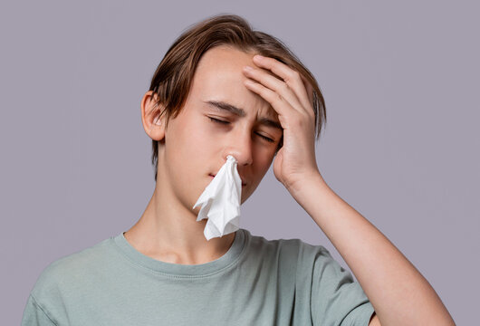 Portrait of unhealthy sick teen boy with napkin close her nose from allergy or rhinitis. Unhappy ill child suffering from running nose, sneezing, blow her nose. Boy having a headache. Headache child