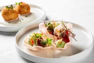 Close-up of gourmet appetizers: crispy shrimp chips topped with avocado mash and garnish in the foreground, and round, breaded croquettes on a creamy sauce in the background.