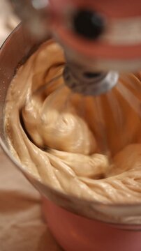 A pink mixer bowl is filled with a thick, creamy batter. The batter is being mixed by a mixer, which is spinning at a high speed. Concept of excitement and anticipation