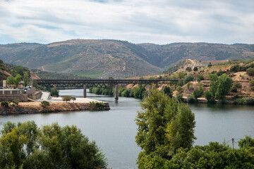 Ponte ferroviária de Barca d'Alva a cruzar o rio Douro, enquadrada por encostas áridas e arborizadas do Vale do Douro Internacional sob um céu parcialmente nublado em Portugal