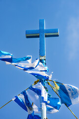 A striking blue cross, covered in glittering material, stands tall against a clear blue sky, surrounded by blue and white prayer flags.