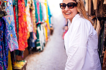 Obraz premium Smiling woman wearing sunglasses, dressed in white jacket, enjoys vibrant market atmosphere, surrounded by colorful textiles and clothing, showcasing joyful shopping experience