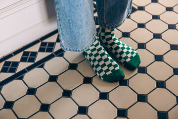 Green and white checkered socks worn by a person with denim jeans, standing on a patterned tiled floor, showcasing a cozy indoor atmosphere with stylish design elements