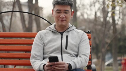 Smiling asian man using mobile phone while sitting on bench in public place. Portrait of cheerful person in glasses and light hoodie wearing modern smartwatch texting message on his smartphone. - Powered by Adobe
