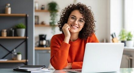 Cheerful young woman with curly hair working at a laptop in a cozy kitchen environment
