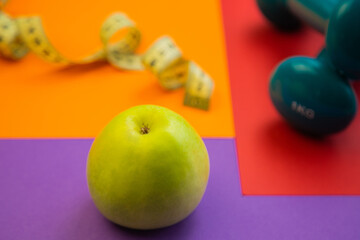 Fresh green apple on purple background, with colorful dumbbell and measuring tape nearby, representing healthy eating and fitness motivation for an active lifestyle