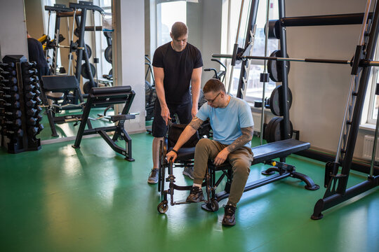 Professional sports trainer assisting hardworking disabled man in wheelchair transition from exercise machine. Physical activity, encouraging inclusive independence, safety and diverse workout