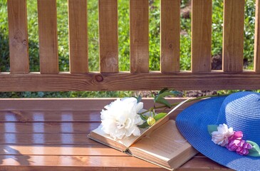 White peony flower on a open book and blue hat on a wooden bench in a summer sunny garden