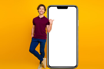 Young curly haired man in a burgundy polo shirt and blue pants presenting a blank white advertising board on a yellow background