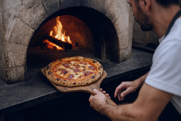 Caucasian young man placing pepperoni pizza in traditional stone oven with burning firewood on a wooden peel in a rustic pizzeria kitchen