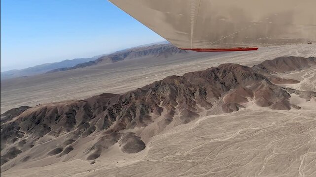 View from a small plane flying over the arid, rugged landscape of the Nazca region, with the aircraft wing visible as it banks near desert mountains before reaching the famous geoglyphs