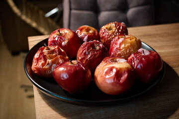 Ripe red apples on a black plate sit on a wooden table, showing varying stages of freshness and decay in a cozy indoor setting