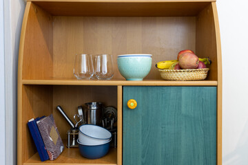 Wooden kitchen shelf with glassware, bowls, utensils, notebooks and fruit basket.