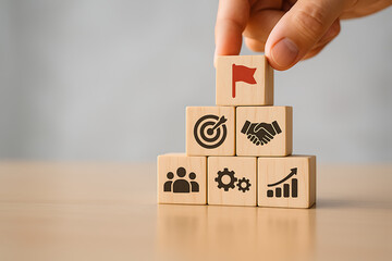 Professional hand placing a wooden block with a red flag icon on top of a pyramid stack symbolizing business objectives teamwork strategy and corporate vision with gray backdrop