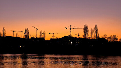 Silhouettes of construction cranes and trees against a vibrant orange-purple sunset.