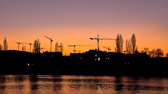 Silhouettes of construction cranes and trees against a vibrant orange-purple sunset.