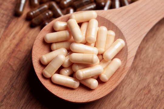 Beige capsules in wooden spoon on table with scattered brown pills