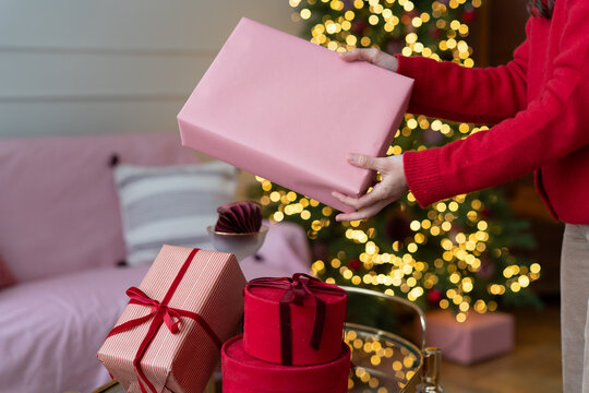 Close-up of a woman standing in front of an illuminated Christmas tree in a living room holding a wrapped Christmas gift