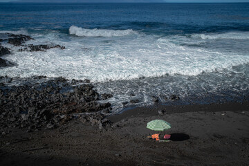 Foamy Atlantic waves meet the rugged black volcanic shore of Tenerife. A green beach umbrella on the dark sand adds a peaceful touch to this dramatic coastal landscape.