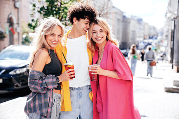 Young three stylish friends posing in the street. Fashion man and two cute female dressed in casual summer clothes. Smiling models having fun. Cheerful women and guy outdoors, Hold and drink lemonade