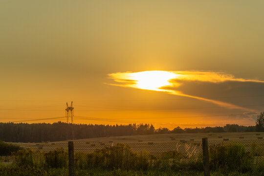 Sunset over harvested field with hay bales and transmission towers in rural landscape. Golden sunset illuminating hay bales in harvested field with wild grasses symbol of countryside tranquility.