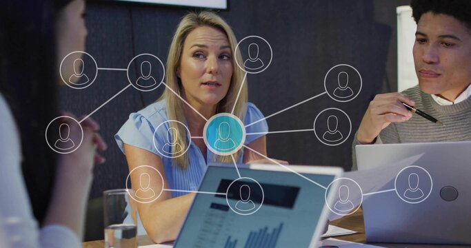 Leading woman wearing blouse speaking to team at wooden table, laptops papers network overlay - Powered by Adobe