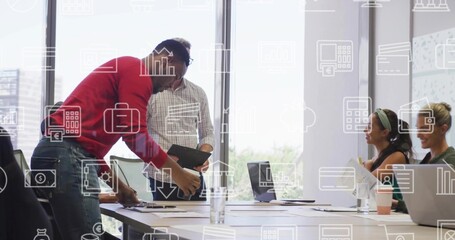 Arranging papers, man in red sweater leaning over office table with laptops, tablet, docs, icons