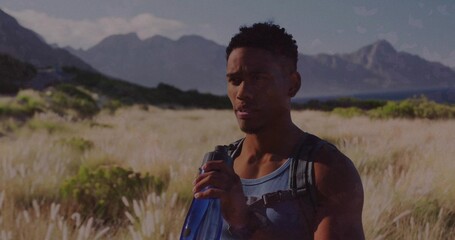 Sipping man pausing in shore meadow holding blue bottle, wearing blue tank top and black backpack