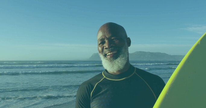 Standing mature man wearing black wetsuit and holding yellow-green surfboard on sandy shore, waves