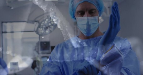 Donning surgeon in blue gown, mask pulling glove on in operating room, with monitors and lamp