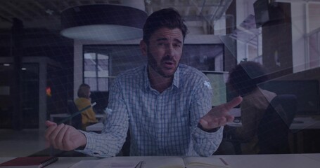 Gesturing man in light blue shirt speaking at open office desk, holding pen and open notebook