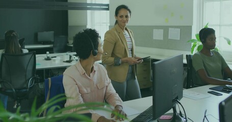 Standing team lead wearing beige blazer holding tablet guiding headset staff in open office