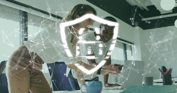 Leaning two business women pointing at silver laptop in office, holographic shield and blue mug