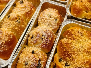 Close-up of golden-brown buns, freshly baked and topped with sweet streusel crumble and raisins, presented in individual foil trays. Perfect for breakfast or dessert.