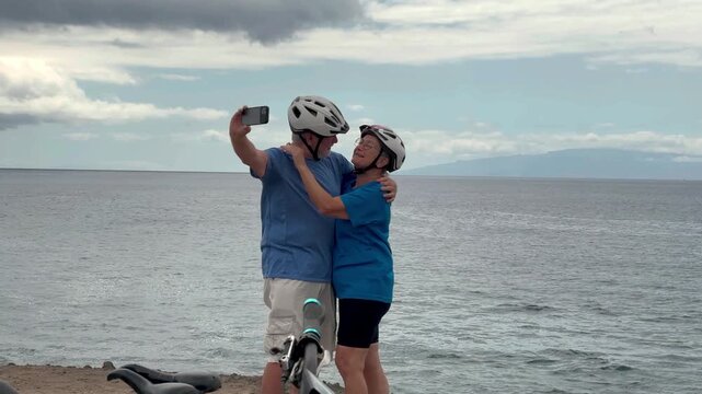 Happy senior couple of cyclists wearing helmets exchange a kiss and take a selfie with phone enjoying riding seaside with electric bicycles. Healthy lifestyle in retirement. Horizon over the sea - Powered by Adobe