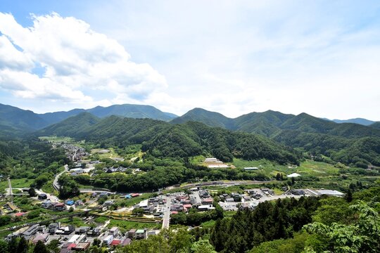 Panoramic summer landscape of a rural Japanese town nestled in lush green mountains, with rice fields, winding roads and houses under a bright blue sky with white clouds.