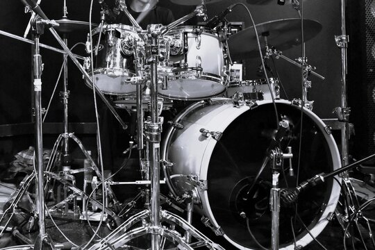 Monochrome close-up of a drum kit on a live music stage, showing detailed hardware, cymbals, microphones and cables, capturing the energy and atmosphere of a concert performance.