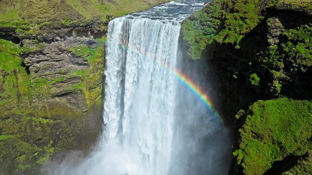 Aerial view of Skogafoss waterfall from a side angle. Drone footage showing dense mist rising from the falls and a rainbow appearing in the sunlight.