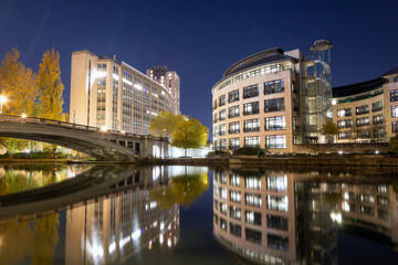 A beautiful night image of two prominent riverside office buildings and an arched bridge, all reflecting brightly in the calm, dark water.