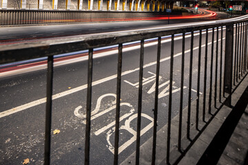 An overhead, diagonal view of a multi-lane roadway with "BUS LANE" marked on the asphalt, seen through a dark metal railing, with blurred light trails of vehicles moving quickly underneath an overpass