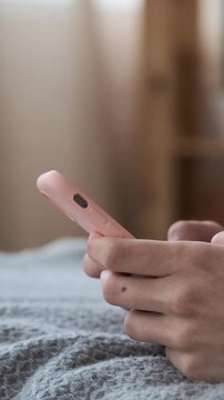 Extreme close-up of female hands holding a pink smartphone while lying on a grey textured blanket in the bedroom, texting or browsing. vertical video